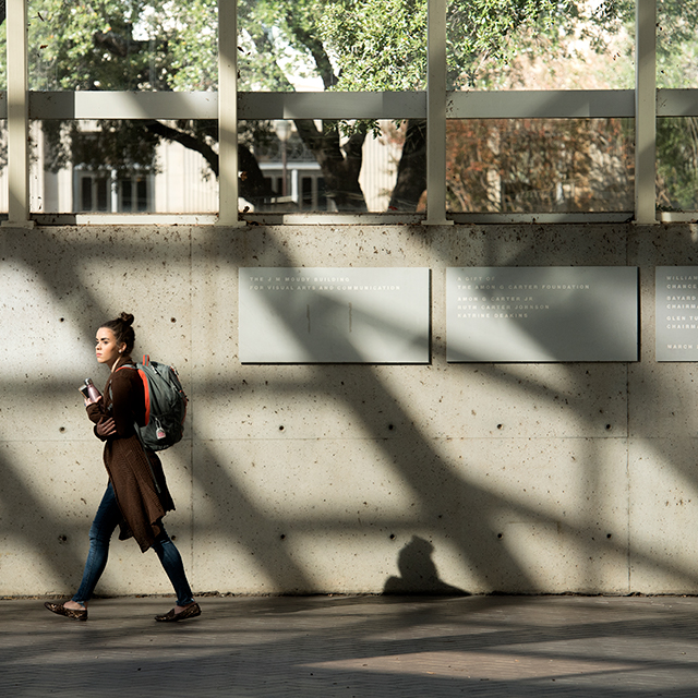 A student carrying a backpack walks through the Moudy Building as the structure's architectural features cast shadows dramatically on a concrete wall
