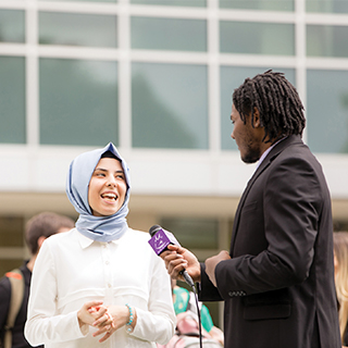 A student reporter holds a microphone up to a student wearing a hijab while a student with a video camera records the interview