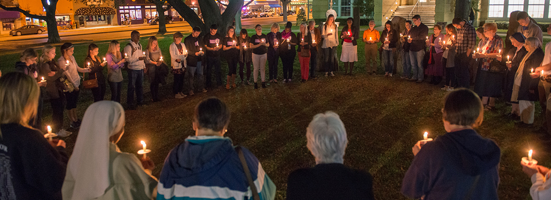 A circle of people holding candles gather for a nighttime prayer vigil on the lawn of the TCU campus.